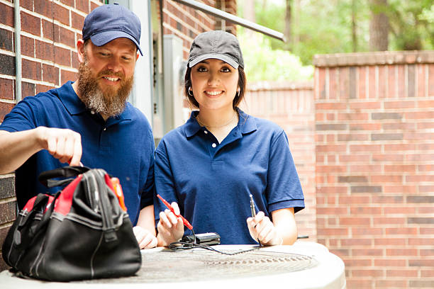 Air conditioner repairmen work on home unit. Multi-ethnic team of one man and one woman repairing a home's air conditioner unit outdoors. They are working on the unit using hand tools in the toolbag.  They wear blue uniforms. technician stock pictures, royalty-free photos & images