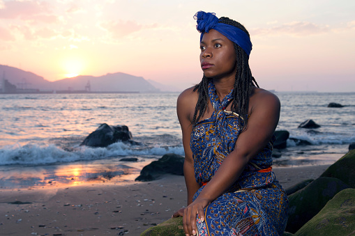 African woman in traditional dress, sitting on a stone at sunset, looking up in a dirty beach.