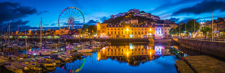 The hotels and villas, quayside shops, pubs and restaurants of Torquay overlooking the harbour illuminated at dusk.