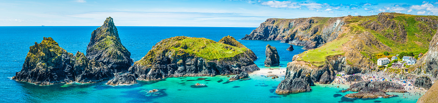 Crowds of tourists enjoying the summer sunshine amongst the dramatic sea stacks and rocky cliffs overlooking the turquoise ocean and idyllic sandy beaches of Kynance Cove, Cornwall, UK.