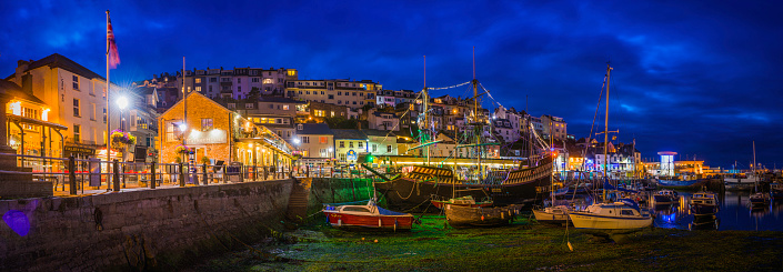 Panoramic view across the still waters of Brixham harbour surrounded by the quaint fishing village cottages illuminated at dusk, Devon, UK.