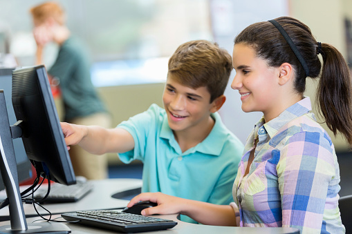 Brunette male and female teenagers using a computer in the school computer lab. Caucasian female high school student sitting at a desk with a mixed race male classmate and working on a computer together. Teenage boy is pointing at computer screen.