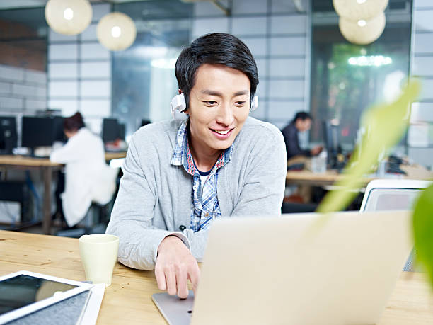 young business person working in office young asian businessman sitting at desk in office working using laptop computer while listening to music with headphone. asian man headphones stock pictures, royalty-free photos & images