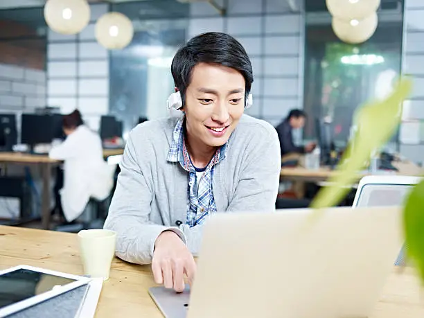 young asian businessman sitting at desk in office working using laptop computer while listening to music with headphone.
