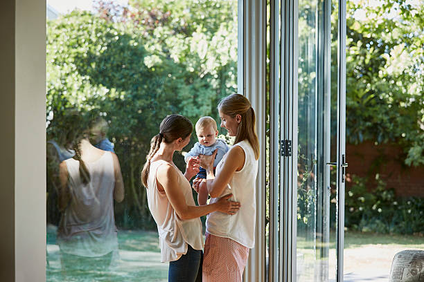 Lesbian couple with baby boy standing by window Happy lesbian couple with baby boy standing by glass window gay long hair stock pictures, royalty-free photos & images
