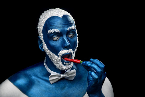 Man painted in blue color with snowy hair and beard eating chili pepper on black background