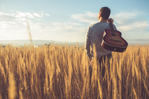 Rear view of a young man standing in the prairie, looking to the sky, carrying a guitar on his shoulder