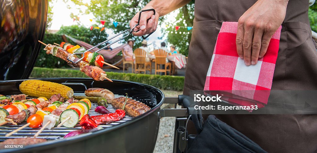 Man grilling meat on garden barbecue party Man grilling meat on garden barbecue party, in the background friends eating and drinking Grilled Stock Photo