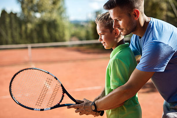 Tennis instructor assisting little boy on the tennis court. Young coach teaching little boy to play tennis on the court during training class. tennis coach stock pictures, royalty-free photos & images