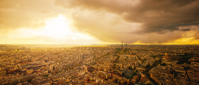 Wide panoramic cityscape of Paris with the Eiffel Tower, Champs de Mars, La Defense, and western Paris at sunset, with dramatic storm clouds over the business district of La Defense.