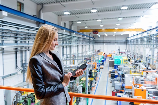 Horizontal color image of blond manager working with digital tablet in large futuristic factory. Blond woman standing on top of a balcony. Focus on businesswoman, futuristic machines in background.
