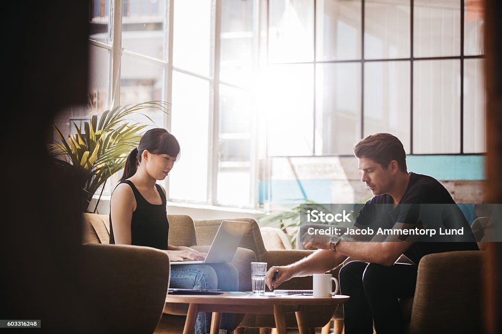 Two businesspeople meeting in lobby area of modern office Shot of two businesspeople meeting in lobby area of modern office. Woman working on laptop with man explaining the project plan. Sofa Stock Photo