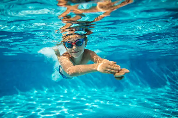 Portrait of smiling little boy enjoying underwater swim in the pool. The boy is gliding by the camera during swimming lesson. Sunny summer day.