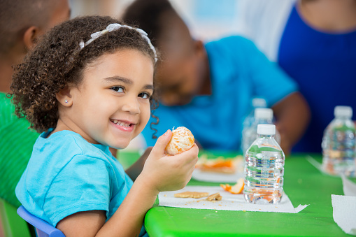 Adorable African American toddler girl eating an orange during lunch time at her preschool. Cute little girl is sitting at a table with preschool classmates while eating an orange for snack time.