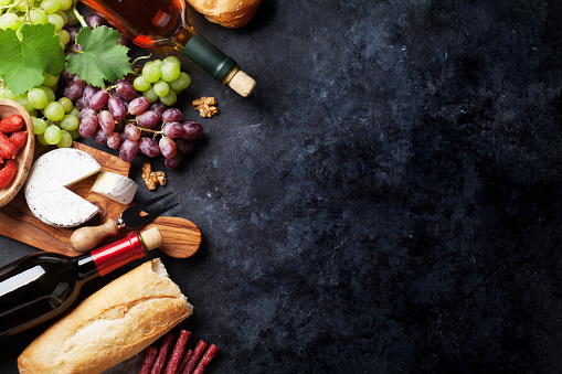Red and white wine bottles, grape, cheese and sausages over stone table. Top view with copy space