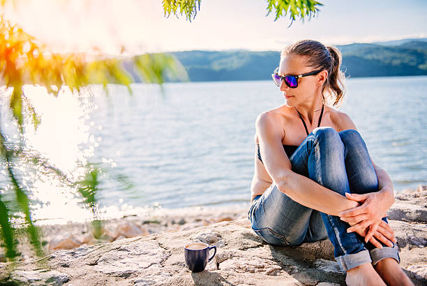 Woman with sunglasses wearing black bikini and jeans sitting on the stone dock and drinking coffee
