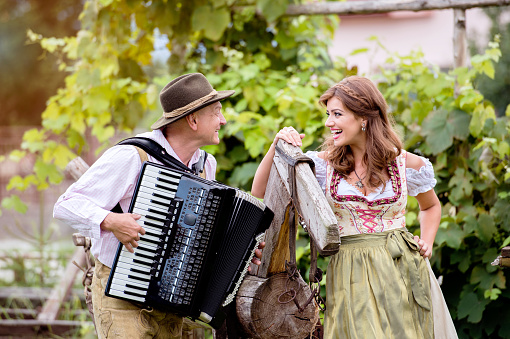 Couple in traditional bavarian clothes standing by an old wooden horse in green garden, man playing accordion. Beer Fest.