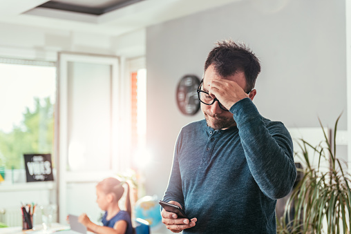 Worried father looking at smart phone and holding hand on forehead, in background his daughter doing homework