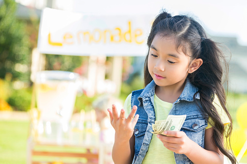 Pretty girl counts on her fingers while holding cash from her lemonade sales. The lemonade stand is in the background.