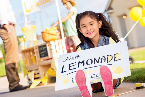 Happy elementary age little girl sits on the ground while holding a homemade 'Lemonade' sign. She is promoting her lemonade stand. Her friend is selling lemonade to a customer in the background.