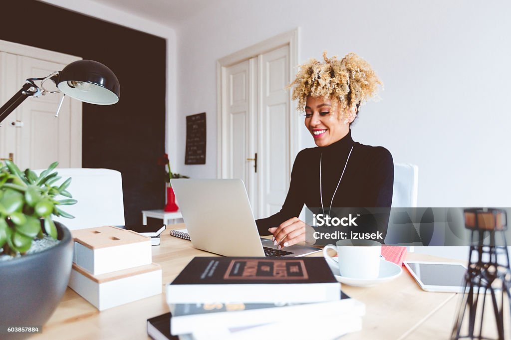 Afro american young woman using laptop in an office Beautiful afro american young woman sitting by the desk in a home office, using laptop. Digital Tablet Stock Photo