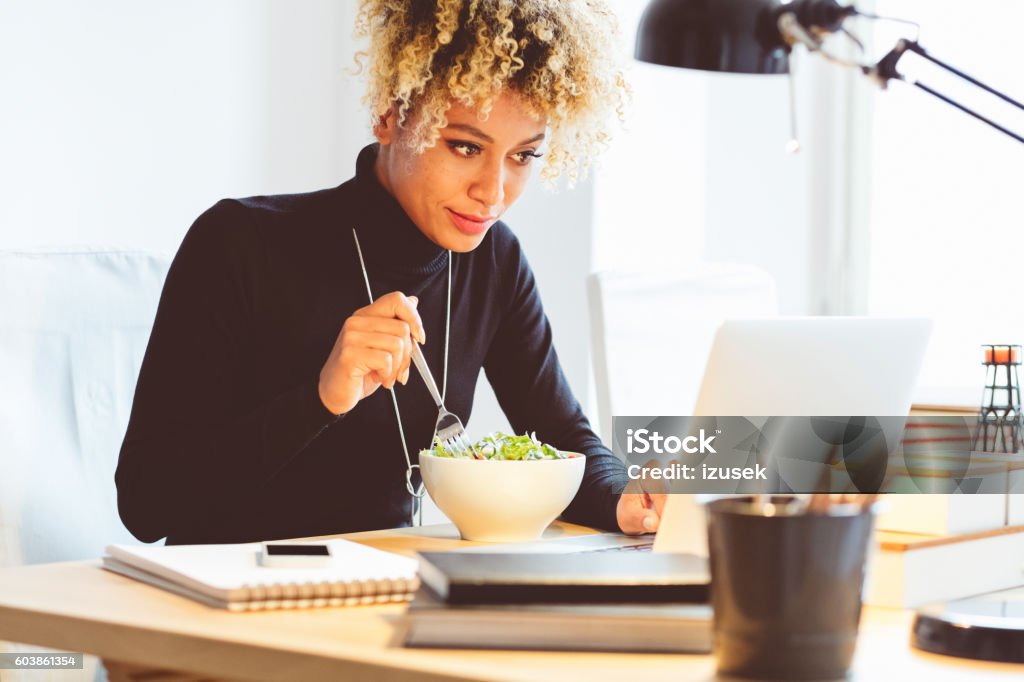 Afro american young woman eating lunch at the desk Beautiful afro american young woman sitting at the desk in an office and eating lunch, using laptop at the same time. Lunch Stock Photo