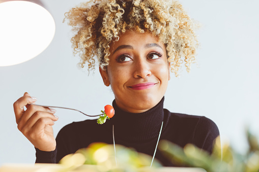 Beautiful afro american young woman eating salad, holding a fork with tomato in hand and smiling. Close up of face.