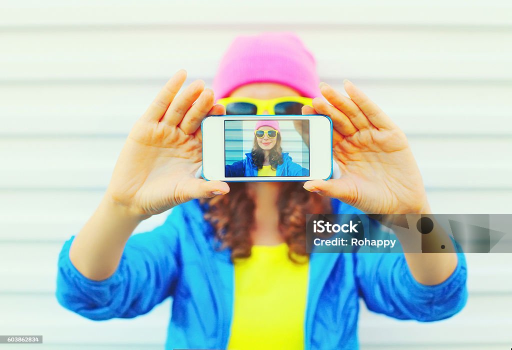 Fashion pretty cool girl taking photo self portrait on smartphone Fashion pretty cool girl taking photo self portrait on smartphone over white background wearing colorful clothes and sunglasses Close-up Stock Photo