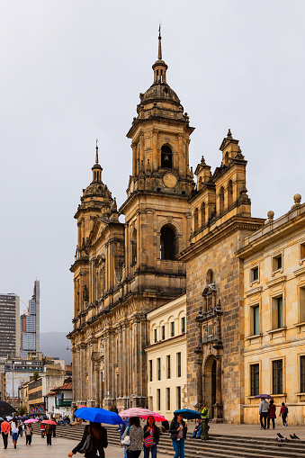 Bogota, Colombia - July 01, 2016: Looking from Plaza Bolivar, in the Andean Capital city of Bogota, in Colombia, South America, to the North Eastern side of the Square. To the right of the image is the Catedral Primada, the seat of the Roman Catholic Archbishop of Colombia. A few yards before the Cathedral, is the Archbishop's chapel. A section of his palace can also be seen. The style of architecture is classical Spanish colonial. In the far background in the haze of the overcast, cloudy sky, the Andes Mountains can be seen.  