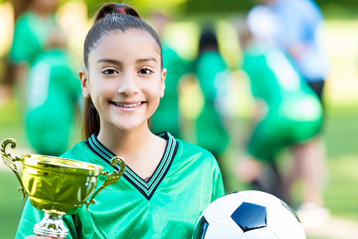 Hispanic female athlete holds golden trophy and soccer ball. She is smiling and wearing a green jersey. Her brown hair is back in a pony tail. She has brown eyes. Her team is blurred in the background.