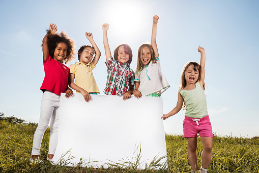 Cheerful children standing in a meadow with hands raised and holding empty placard. Copy space.