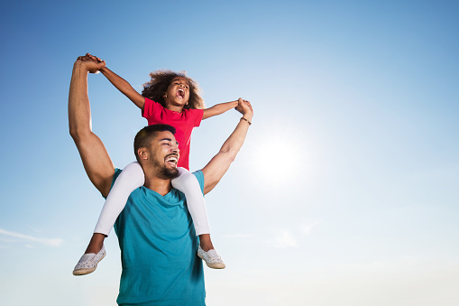 Low angle view of cheerful African American father carrying his daughter on shoulders against the sky while holding hands and shouting. Copy space.
