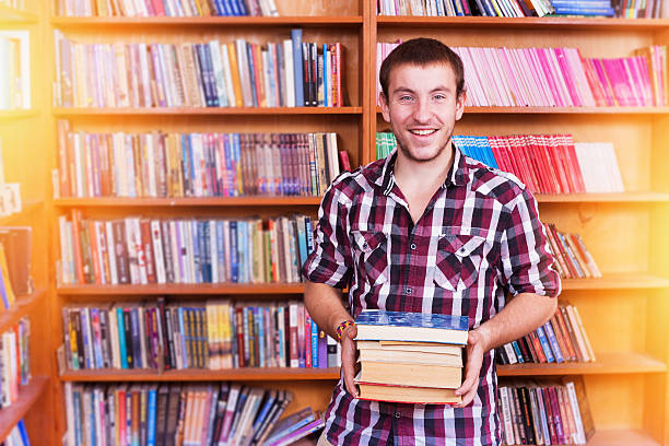 istock Attractive young man holding stack of books in the library 603998958
