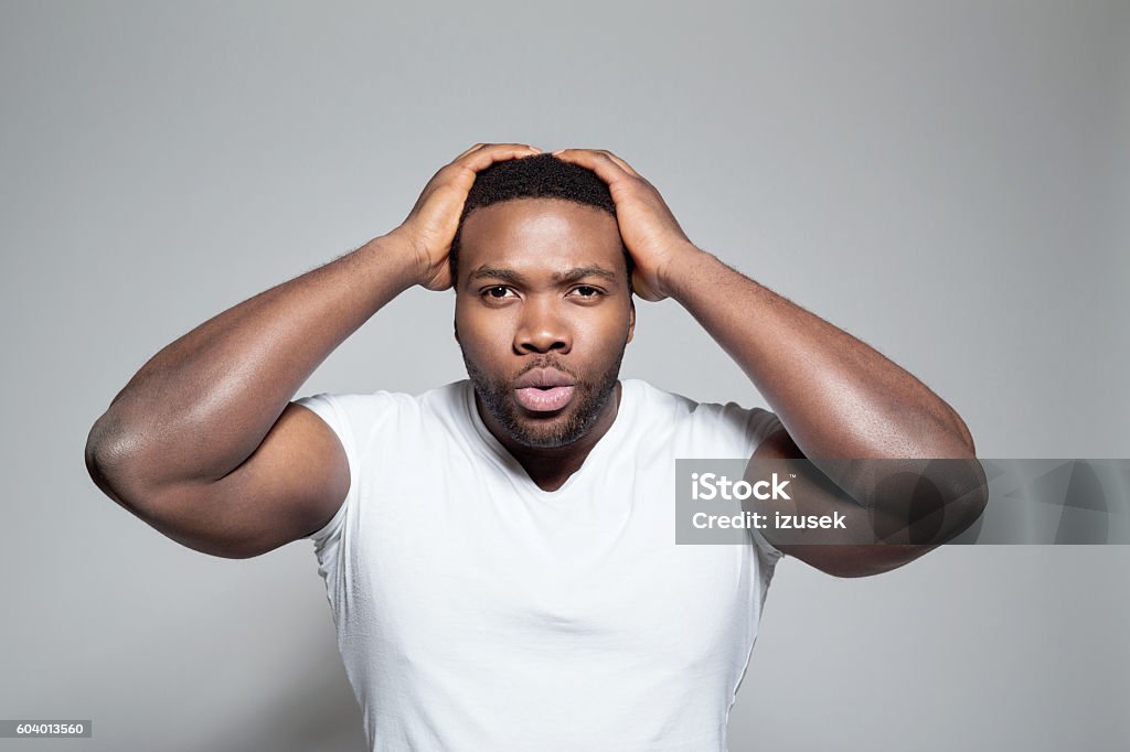 Portrait of surprised afro american young man Portrait of afro american young man wearing white t-shirt, standing against grey background with raised arms, staring at camera. African-American Ethnicity Stock Photo