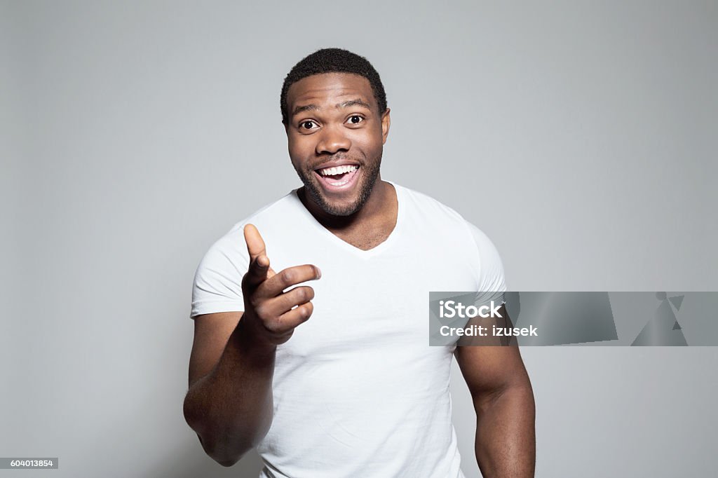 Portrait of excited afro american young man Portrait of excited afro american young man wearing white t-shirt, standing against grey background, pointing at camera with index finger. Body Building Stock Photo