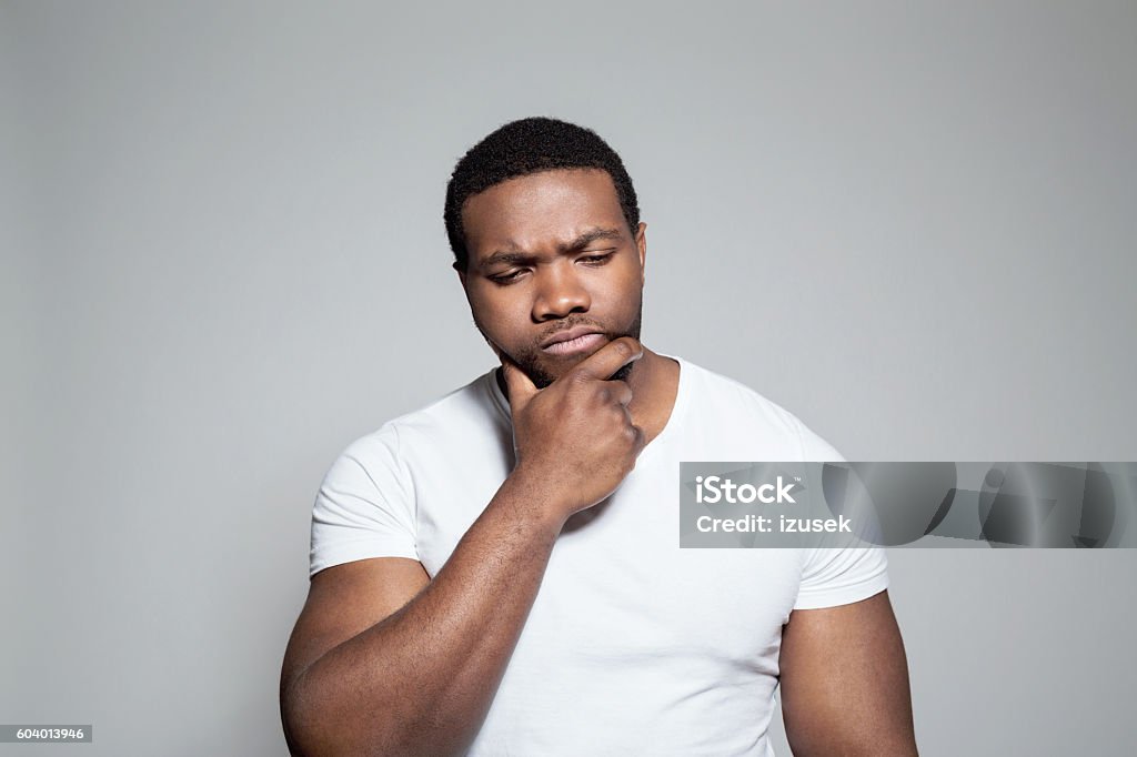 Portrait of worried afro american young man Portrait of pensive afro american young man wearing white t-shirt, standing against grey background with hand on chin. Body Building Stock Photo