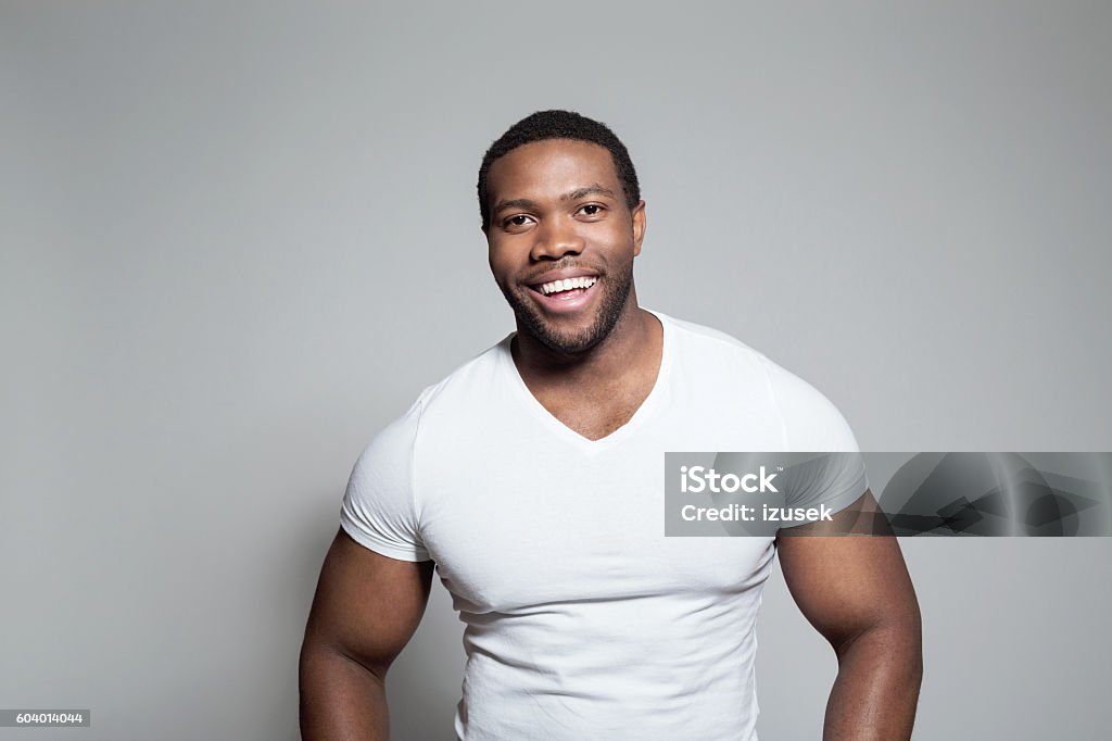 Portrait of happy afro american young man Portrait of happy afro american young man wearing white t-shirt, standing against grey background, laughing at camera. African Ethnicity Stock Photo