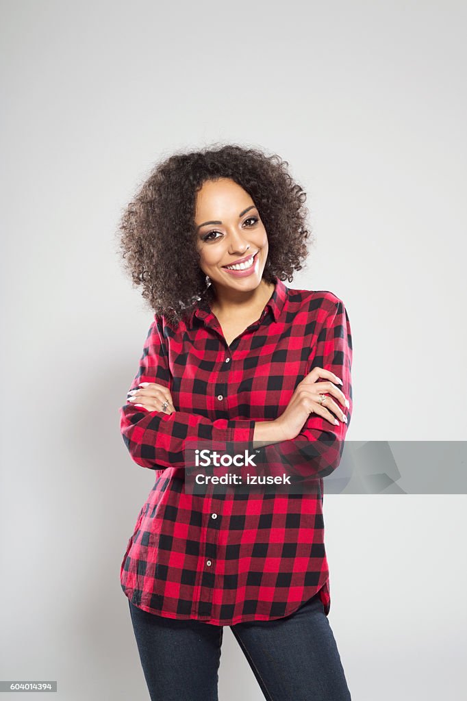 Portrait of happy afro american young woman Portrait of cheerful afro american young woman wearing casual checkered dress, standing against grey background with arms crossed, laughing at camera. Adult Stock Photo