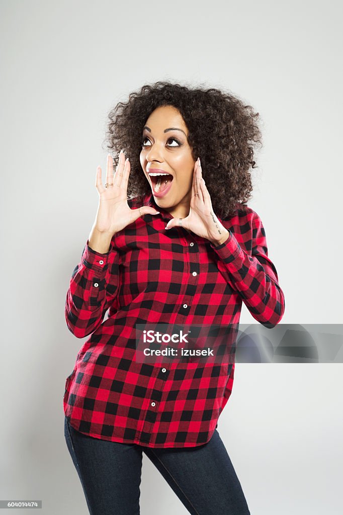 Excited afro american young woman shouting Portrait of excited afro american young woman wearing casual checkered dress, standing against grey background and shouting. Adult Stock Photo