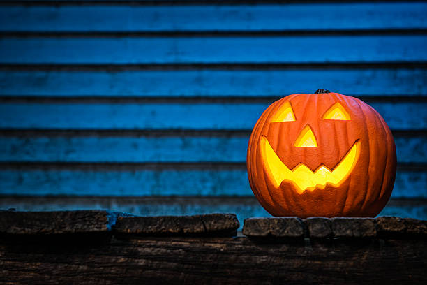 A pumpkin carved with a big smile on this Jack O' Lantern's face, looking at the camera, sitting on an wooden porch at night illuminated by a blue moon light with old dirty house siding in the background, just in time for Halloween.