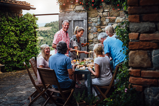 A group of mature friends are sitting around an outdoor dining table, eating and drinking. They are all talking happily and enjoying each others company. The image has been taken in Tuscany, Italy.