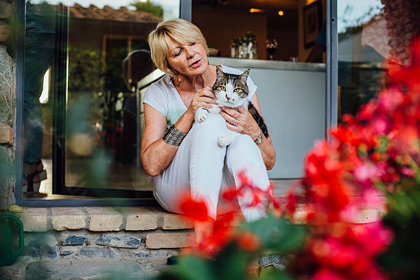 A mature woman is sitting outside of her home with her pet cat on her knee. The shot is framed by a beautiful plant with red flowers.