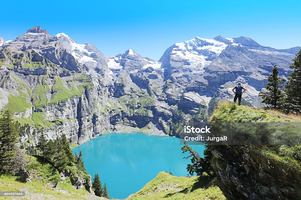 Hiker Admiring Oeschinen Lake in Switzerland Hiker is standing at the edge of a mountain looking at turquoise Oeschinensee below. This picture is taken from the Heuberg - Oberbergli - Oeschinensee hiking trail at the UNESCO World Heritage Site near Kandersteg in Berner Oberland region in Switzerland. Lake Oeschinensee Stock Photo