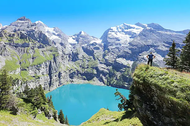 Hiker is standing at the edge of a mountain looking at turquoise Oeschinensee below. This picture is taken from the Heuberg - Oberbergli - Oeschinensee hiking trail at the UNESCO World Heritage Site near Kandersteg in Berner Oberland region in Switzerland.