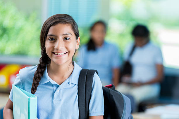 Confident Hispanic middle schoolgirl before class Cheerful Hispanic private middle school student smiles before class. She is standing in her classroom. He has a braid and is wearing a backpack and a school uniform. Students are in the background talking. uniform stock pictures, royalty-free photos & images