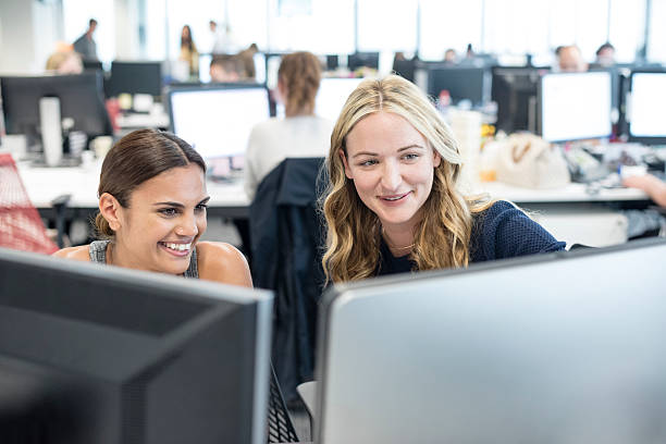 zwei frauen, die im büro arbeiten und auf den computer schauen, lächelnd - minority professional occupation business ethnic stock-fotos und bilder