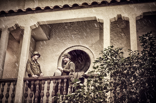 Two WWII US Infantry Soldiers Standing On A Balcony Talking.  It is raining and they seem relaxed and maybe enjoying a little R&R.