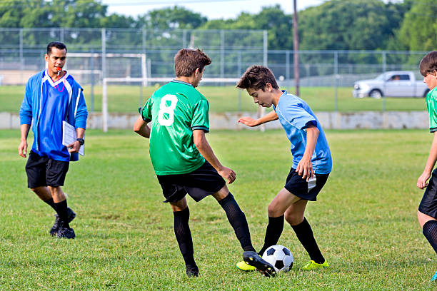 Two teenage male soccer players face off against one another. One boy is wearing a green jersey and the other is wearing a blue jersey. They are trying to take possession of the ball. A mid adult male coach is in the background. He is wearing a blue hoodie. A soccer goal and fence are in the background along with a truck parked in the parking lot. They are playing the game in an athletic complex.