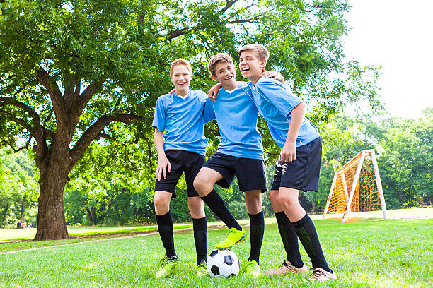 A group three teenage boy soccer players kid around before or after their soccer game. They are all laughing and being silly. They are wearing light blue jerseys with black shorts and black socks. The soccer field and a soccer goal are in the background.