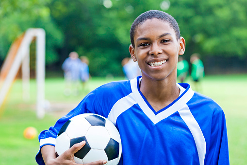 Handsome African American teenage soccer player holds a ball and smiles after winning the game. He is looking at the camera and is wearing a blue jersey with white stripe. His teammates and opposing team members are in the background along with the soccer goal.